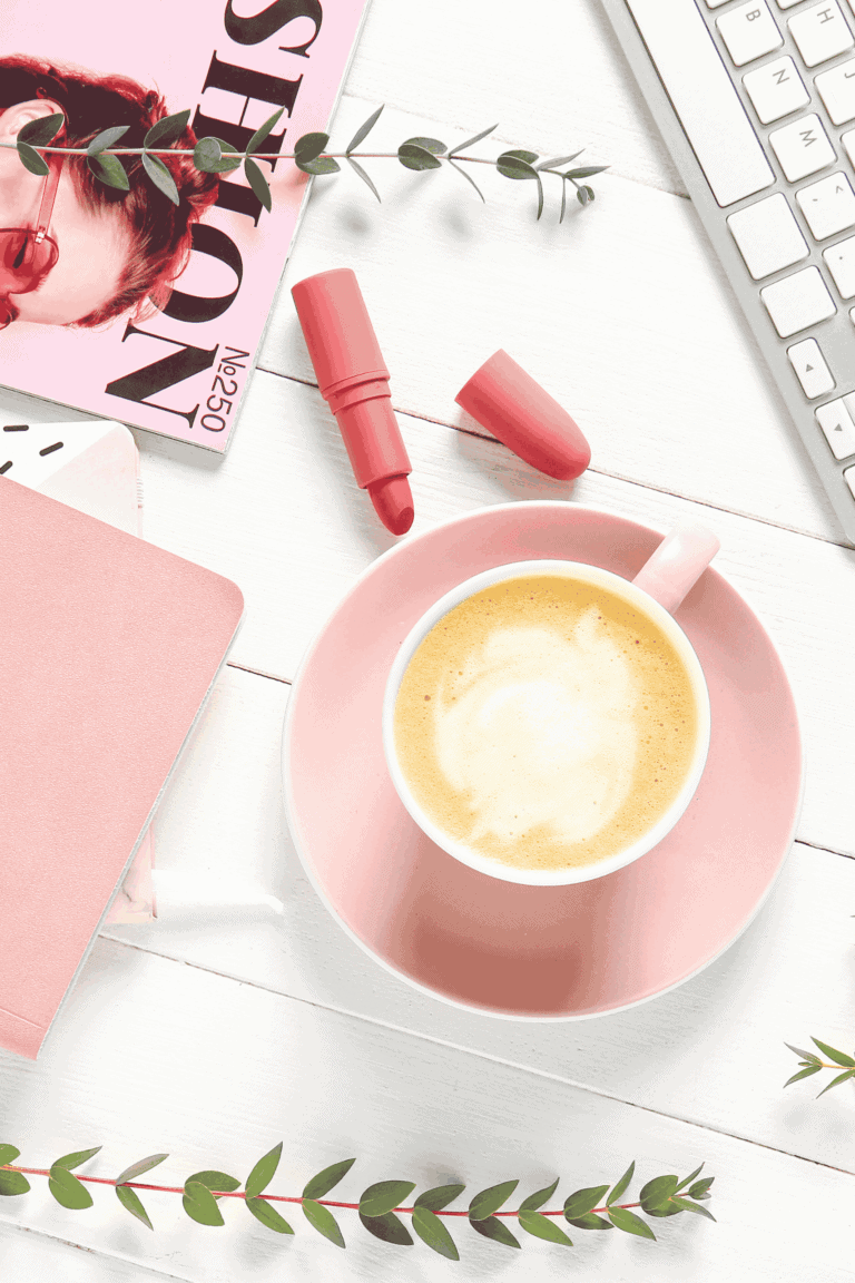 Flat lay of coffee in a pink cup and saucer with a fashion magazine, computer keyboard, pink journal and pink lipstick on a table