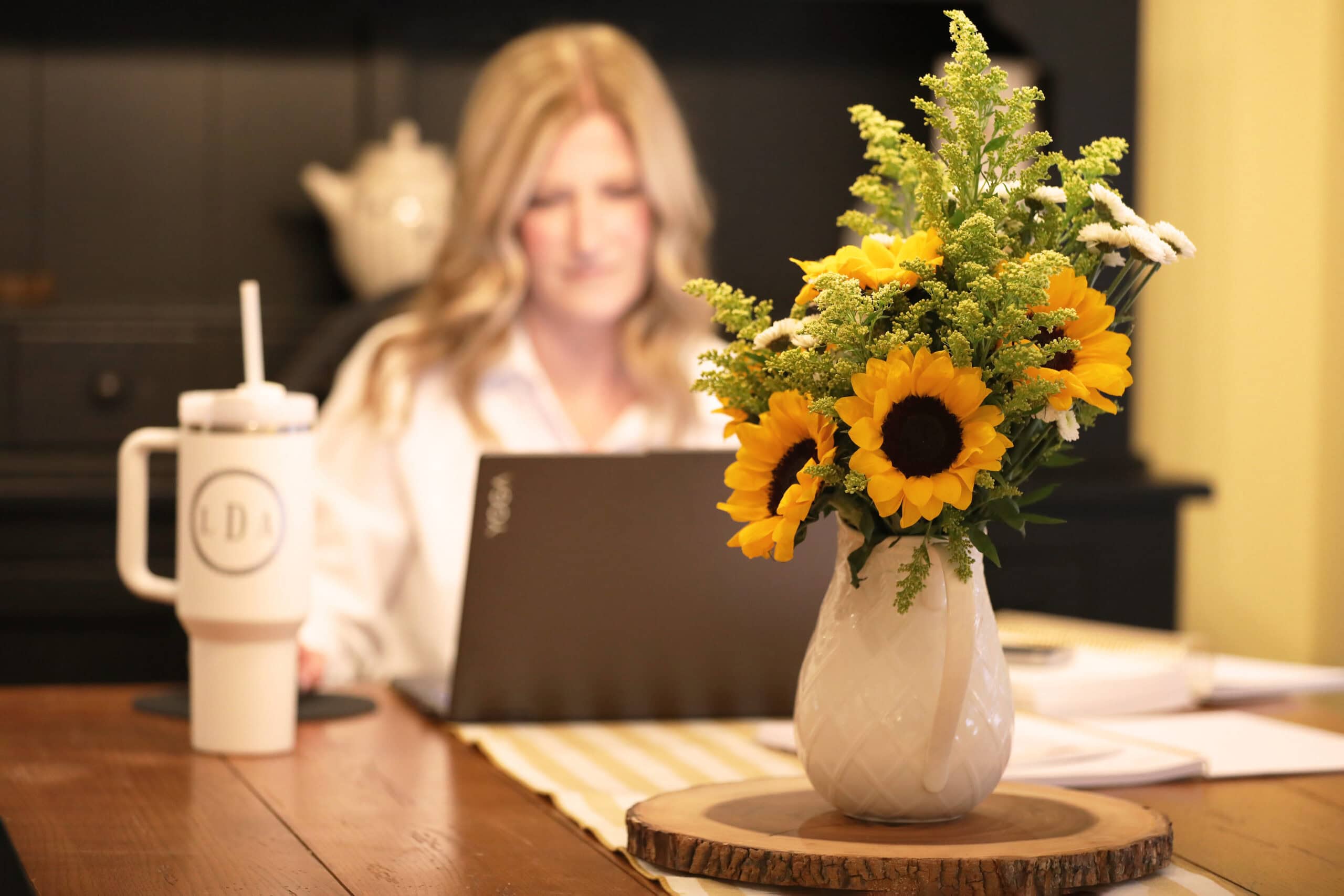 photo of pitcher with sunflowers on table with blurred background of woman sitting looking at her laptop computer.
