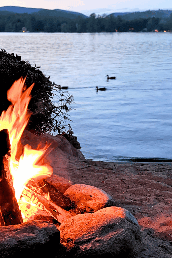 Photo of crackling campfire just before dusk with a quiet lake and mountainview directly behind. A family of ducks is swimming by in the lake beyond the fire.