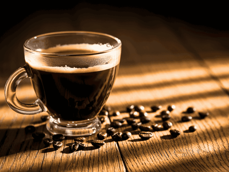 a close-up photo of hot black coffee in a clear coffee mug on a wooden table with coffee beans strewn about on the table next to it in the morning sunlight