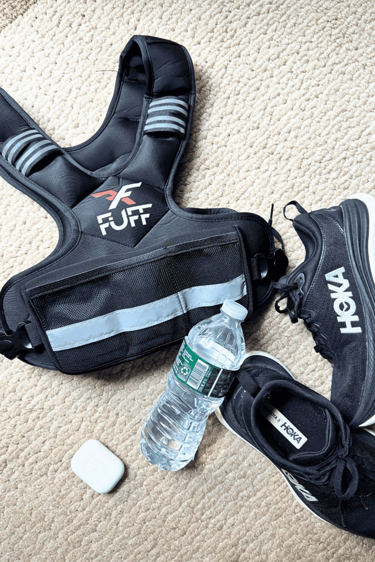 Flatlay photo of a black weighted vest, pair of black Hoka sneakers, a water bottle and an airpods case