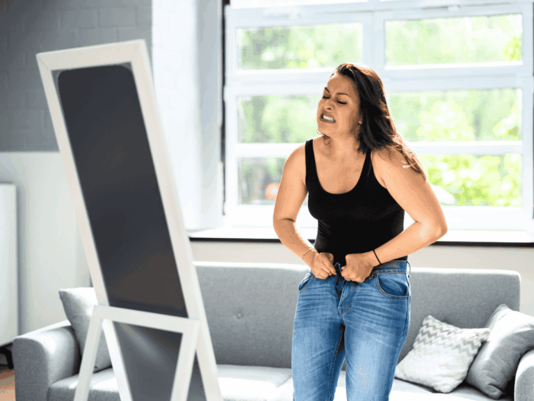 photo of a frustrated looking woman with dark hair looking in the mirror while struggling to put a pair of jeans on.