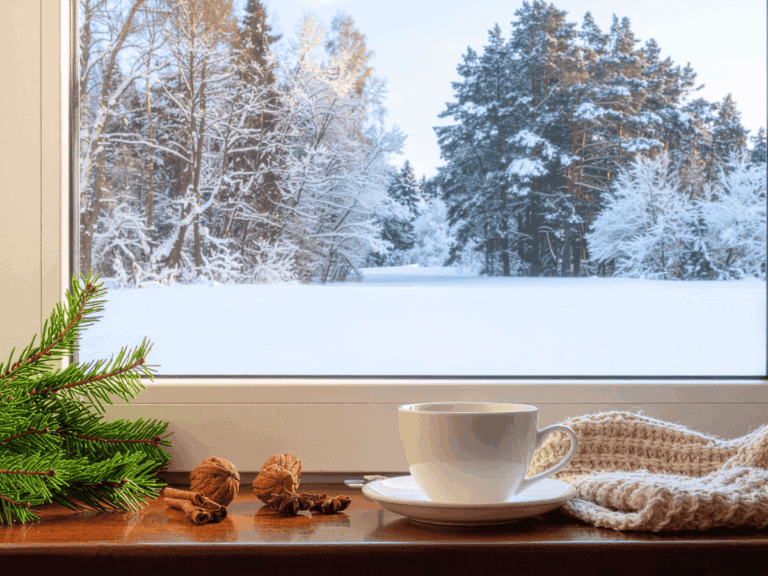 Image of window and sill with a white cup of coffee on sill alog with pine sprig and chestnuts, looking out on a serene snowy scene with pine trees covered in white snow.