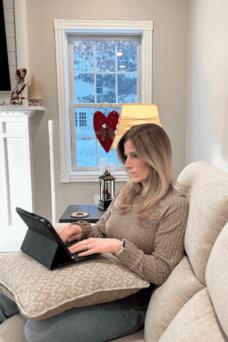 Profile of woman with long brown and blond hair, sitting on a beige couch typing on her ipad. There is a lamp glowing beside her and light gray walls and white trimmed window behind her looking out on a wintry scape.