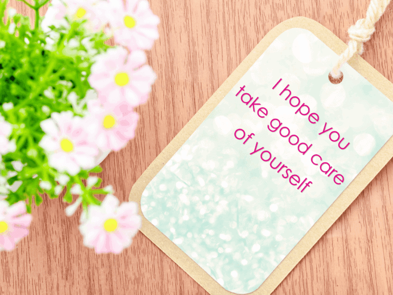 Tag with the words “I hope you take good care of yourself” resting on a wooden surface beside soft pink flowers.
