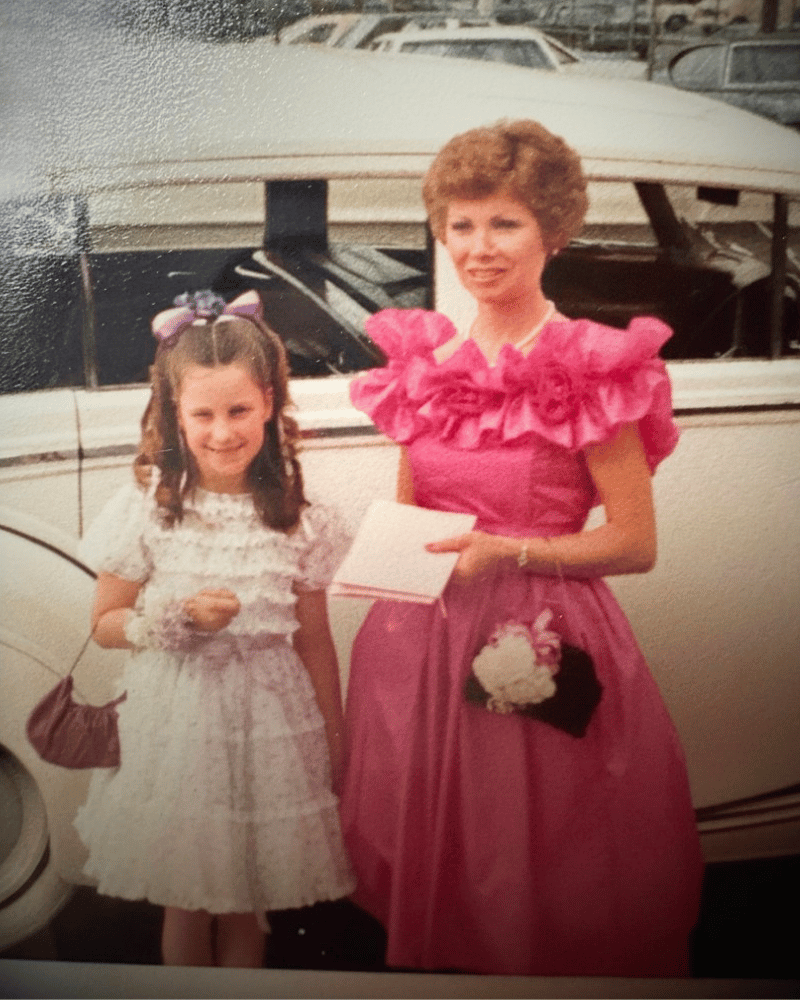 Mother and daughter in elegant dresses standing next to a classic car.