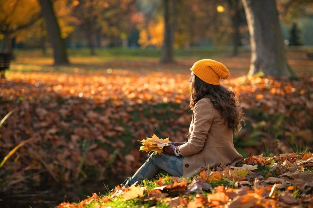 Woman sitting on fallen leaves in autumn park, enjoying peaceful moment.