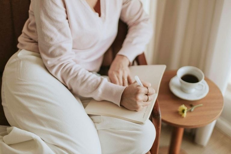 Woman writing in a notebook with coffee on a side table.
